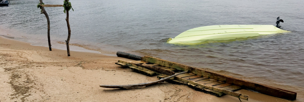 Pale green overturned boat in water near beach with sticks.
