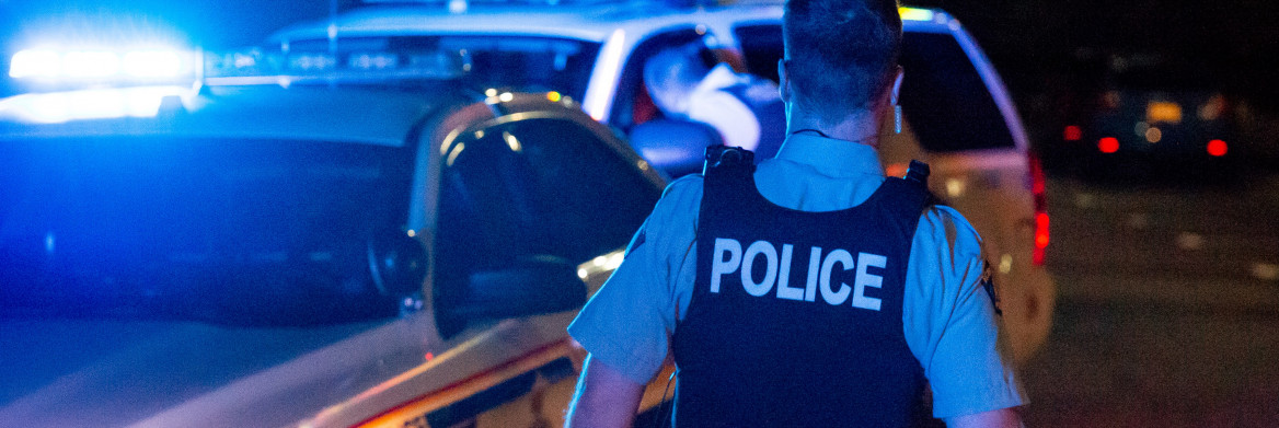 A male RCMP officer approaching a police car at night.