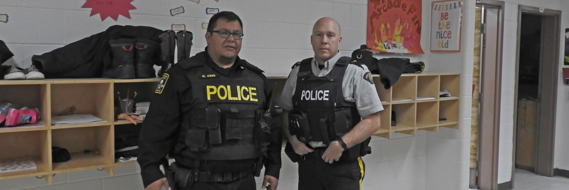 Two male police officers stand next to a wall-mounted storage cubicle in a school.