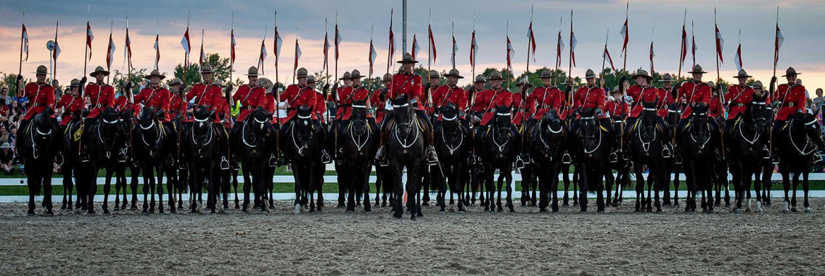 A line of RCMP officers in red serge ride black horses while each holding red and white lance pennon.