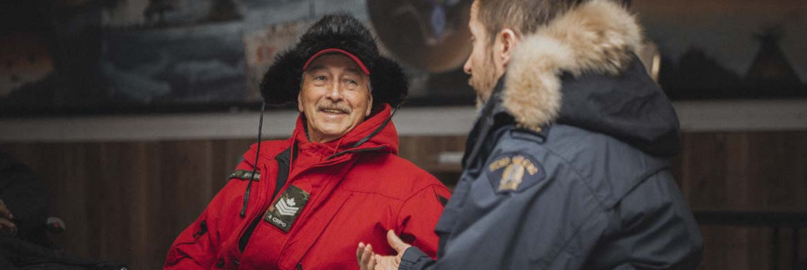 Un homme souriant portant un parka rouge et un chapeau de fourrure est assis face à un agent de la GRC portant un parka.