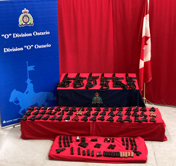 Seized firearms and high capacity magazines on display with RCMP O Division banner and Canadian flag in background. The RCMP Cornwall Border Integrity Unit seized 53 restricted pistols, 6 prohibited rifles, and over 100 prohibited high capacity magazines.