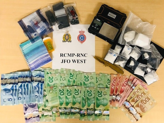 A quantity of cash with denominations between $5 -$100 bills, a number of ziplock bags of cocaine, a knife, a small set of digital weigh scales and other items consistent with drug trafficking is displayed on a table with a RCMP-RNC JFO West poster.