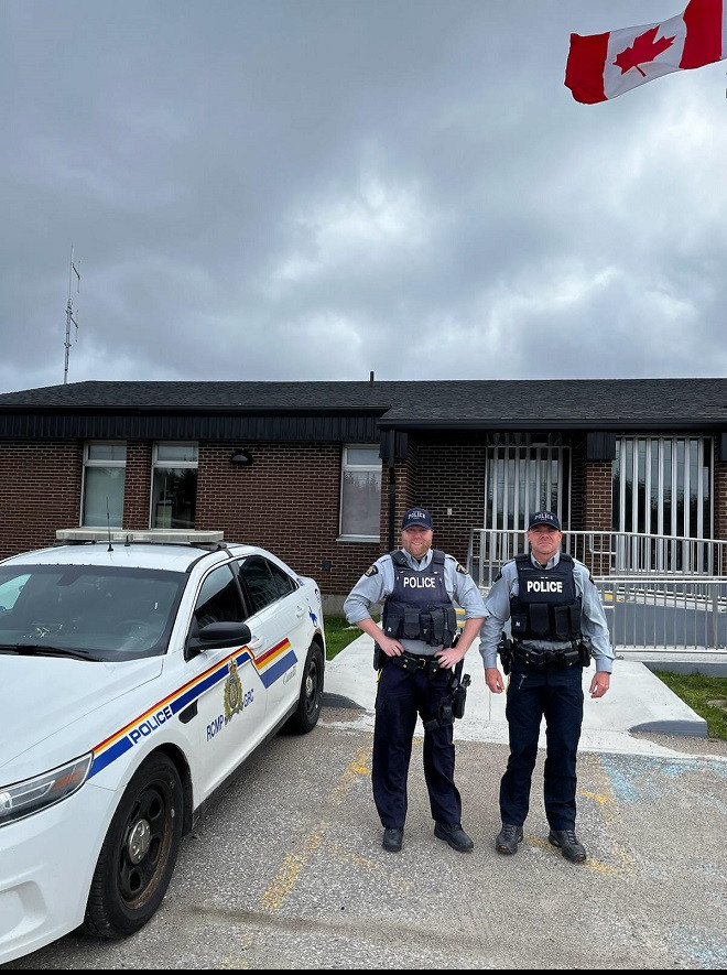 Two RCMP officers in uniform stand in front of Port aux Basques RCMP detachment between a Canadian Flag and a marked RCMP police cruiser.
