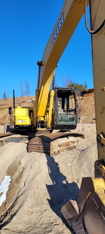 Une excavatrice John Deere jaune est stationnée dans une carrière de sable, la benne sur le sol. Le ciel bleu figure à l'arrière-plan.