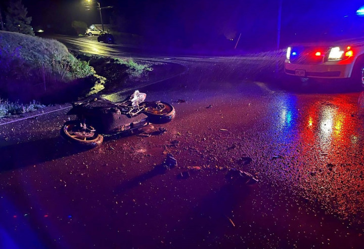 Une motocyclette renversée sur la chaussée, le soir, avec des débris ici et là sur la chaussée. Un véhicule de police identifié avec ses gyrophares activés est visible dans le coin supérieur gauche.