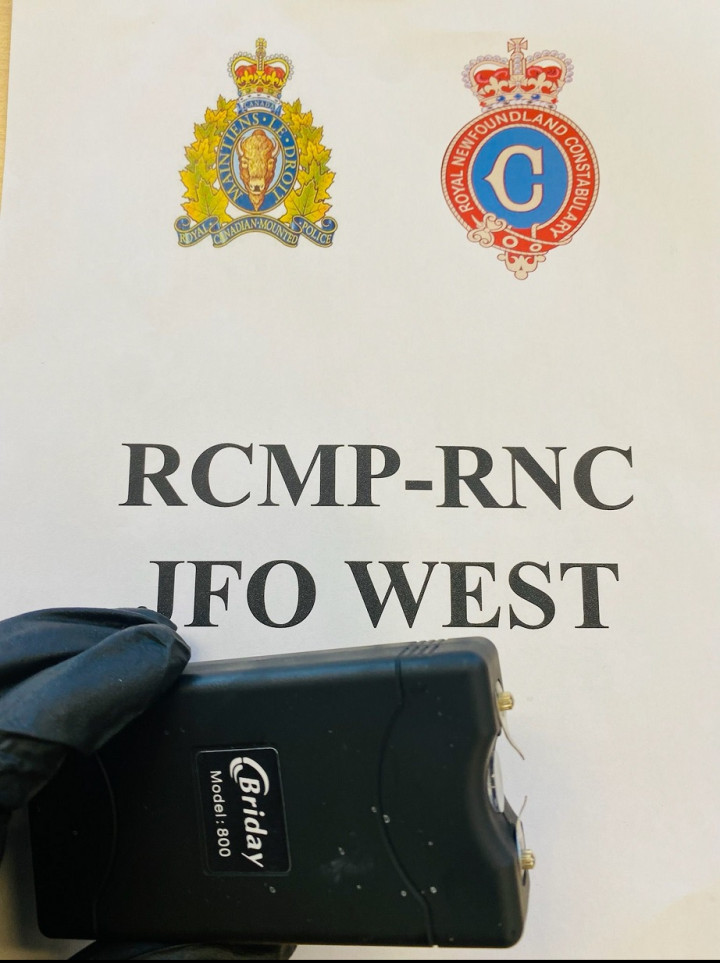 Arme à impulsions noire rectangulaire de marque Briday, modèle 800, devant une affiche du GAIO de la GRC-RNC sur laquelle on peut voir les emblèmes de deux services de police.