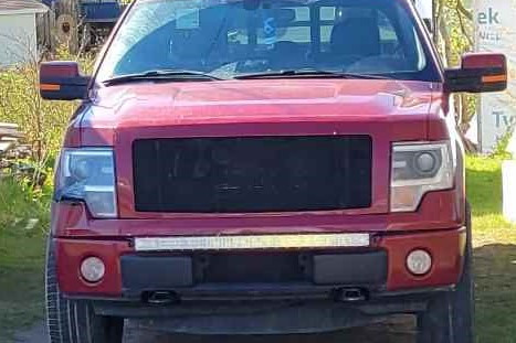 Vue du devant d'une camionnette rouge, avec la calandre noircie. Une remorque est attachée derrière la camionnette. Des remises et une maison sont visibles en arrière-plan.
