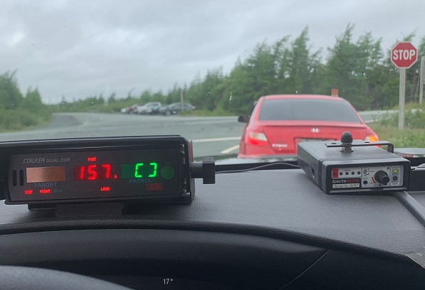 Un radar de police installé sur le tableau de bord d'une voiture de police affiche une vitesse de 157 km/h. Une voiture rouge de marque Honda est rangée en bordure de l'autoroute.