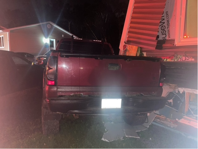 The rear-end of a burgundy pick-up truck is observed. The passenger side of the truck has contact with a home and the driver side of the truck has contact with a parked dark colored SUV.