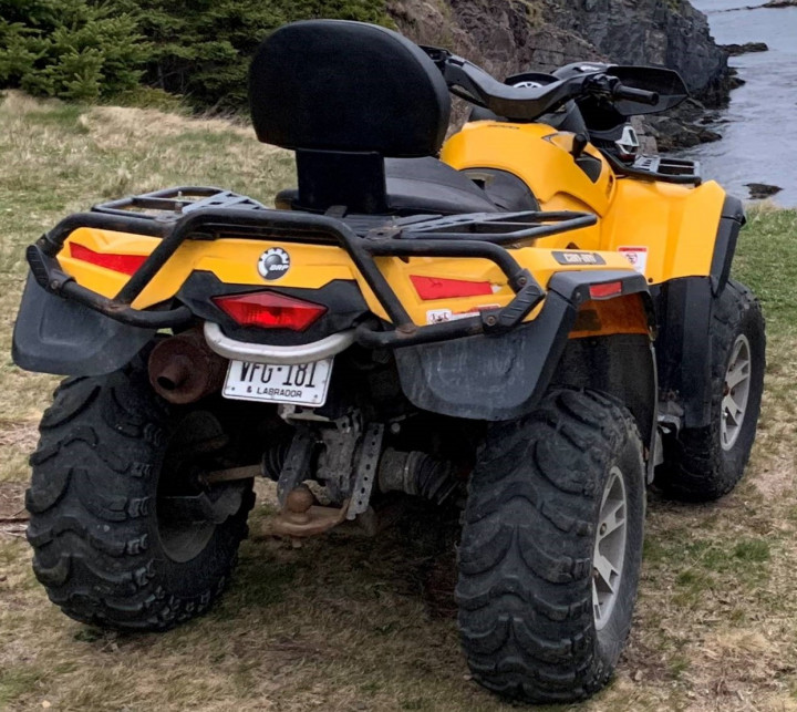 A black and yellow all-terrain vehicle sits on a grassy cliff, facing the water. The license plate, reading 'VFG 181' is visible on the back of the vehicle. 