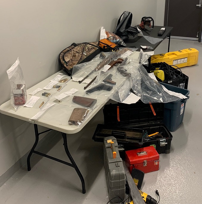 A number of firearms, ammunition, parts of firearms and cases are displayed on a table inside a building. A number of tools are displayed on the floor in front of the table.