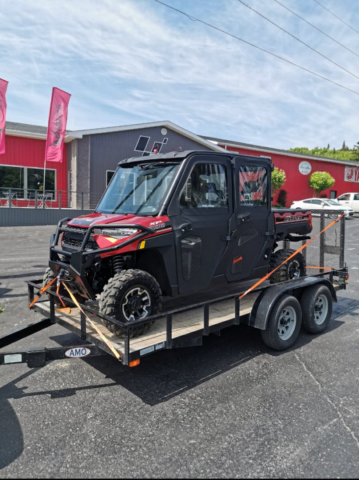A red and black four-door side-by-side Polaris Ranger XP off-road vehicle is pictured on a small utility trailer. A commercial building is in the background.