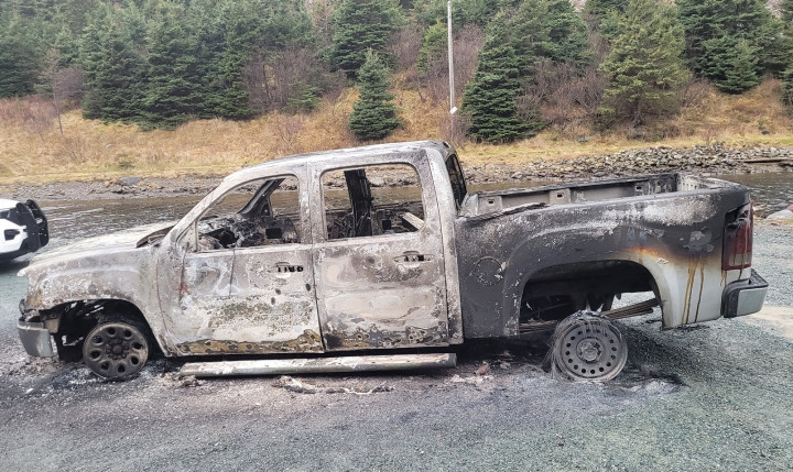 A four-door GMC pickup truck rests on its rims heavily damaged by fire. It is parked on a gravel surface in front of some water and a wooded area. 