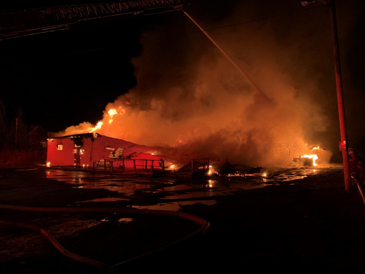 A building is engulfed in flames at night. Water is being deployed overtop the building from the ladder of a fire truck.