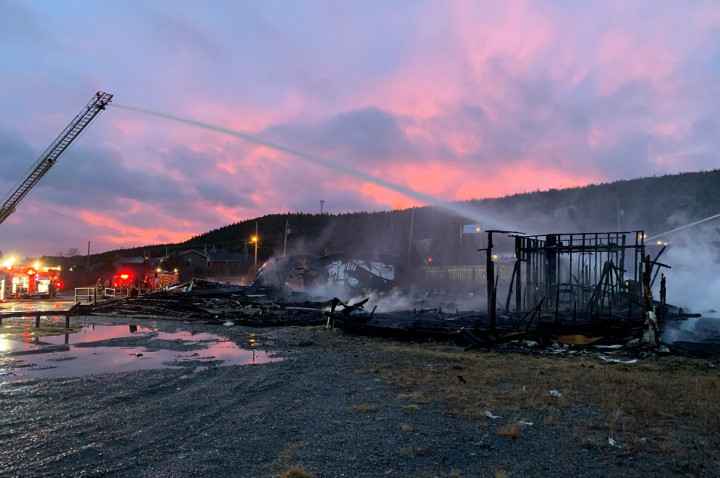 A building is completely destroyed by fire. Water is being sprayed on the remaining structure. Fire trucks are visible in the background.