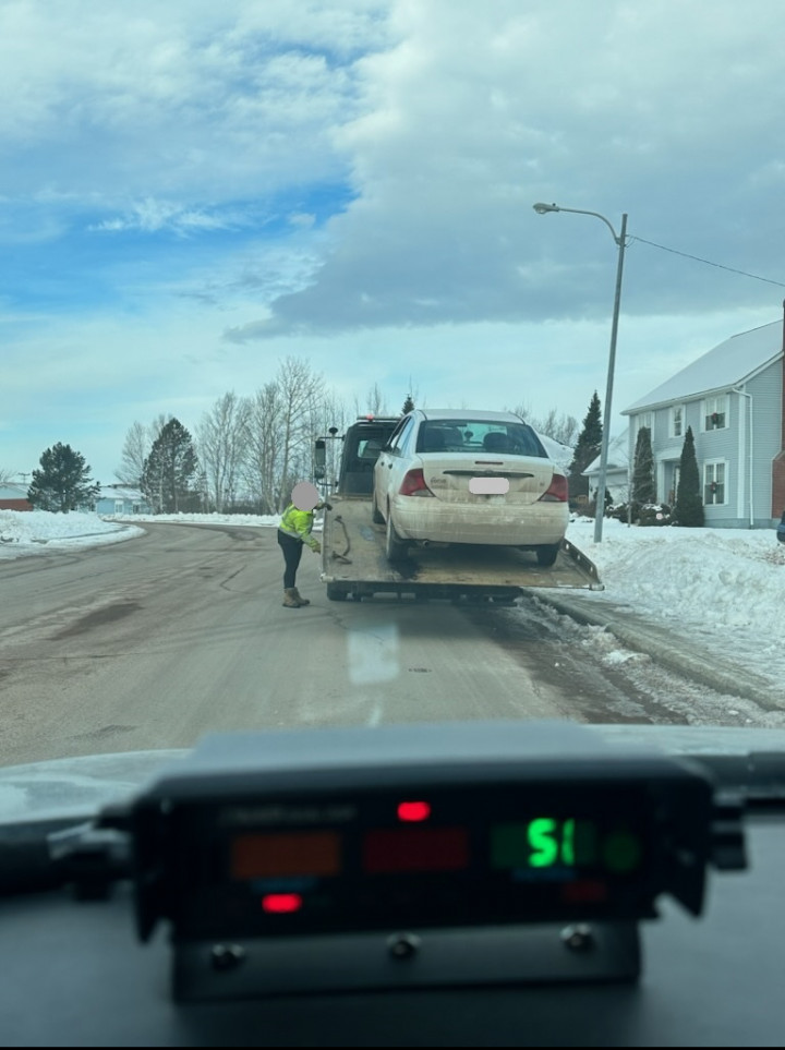 A picture is taken from inside a police vehicle on a cloudy day. A radar set is mounted on the dash of the patrol car. Outside and in front of the police vehicle, a white car is being loaded onto a tow truck.