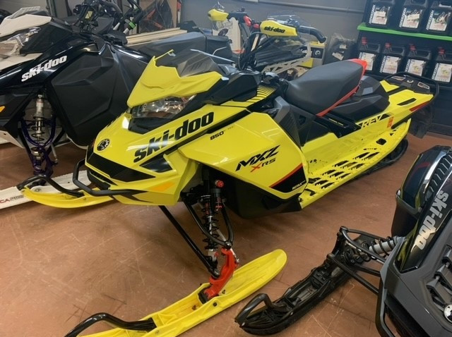 A 2020 yellow and black Ski-doo MXZ XRS snowmobile is displayed inside a building, along side other snowmobiles. 