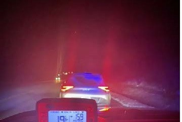 A radar mounted on the inside of a police vehicle displays a speed of 159 km/h. A car is parked in front of the police car on the side of a highway at night.