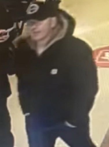 On voit un homme de race blanche dans un magasin. Il porte une casquette de baseball noire et un manteau d'hiver à capuchon noir.