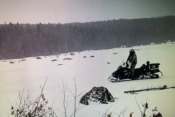 A black and white image of a person on a snowmobile on a snow-covered open area. Trees are observed in the background.