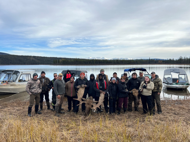 Des participants, souriants, à la chasse scolaire de l'école secondaire de Watson Lake se tiennent devant un lac. Deux élèves soutiennent la tête d'un orignal abattu se trouvant sur le sol devant eux. Quatre bateaux sont sur le lac, derrière eux, avec des montagnes aux sommets enneigés au loin. 