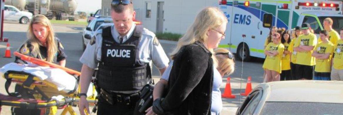 Des élèves assistant à une simulation d'un accident de voiture observent une policière menotter une femme près d'un véhicule. Un policier et une ambulancière sont aussi sur les lieux.