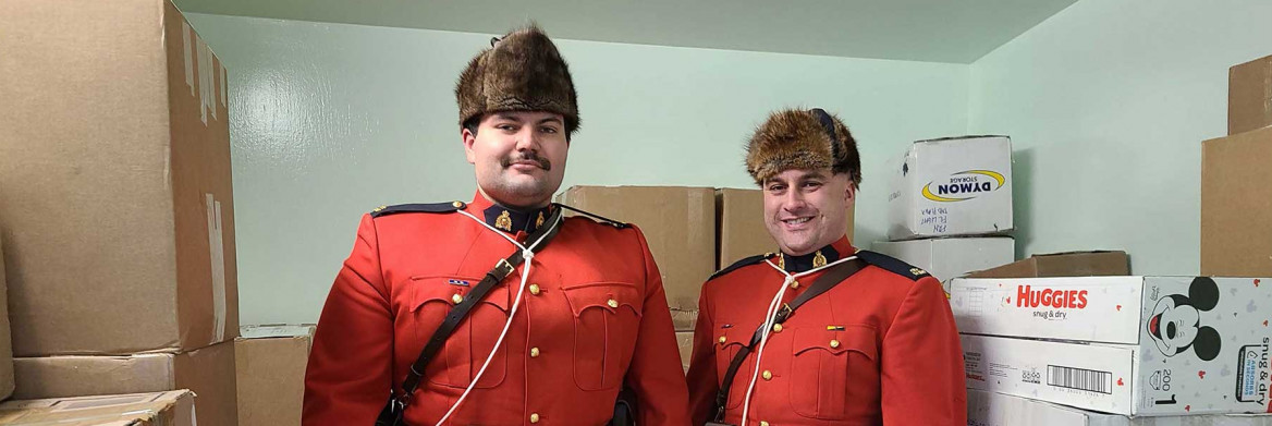 Two RCMP officers in red serge stand in a detachment cell lined with boxes full of donated items.