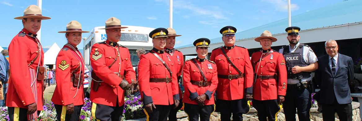 Ten RCMP employees stand in front of the Canada, Métis, and Saskatchewan flags. Eight are wearing red serge, one is wearing a duty uniform and one is wearing a suit.