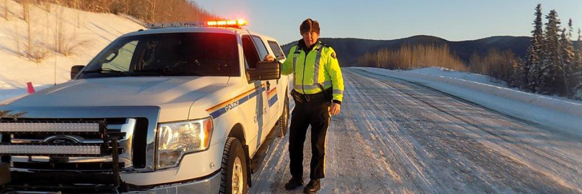 Male RCMP officer in a reflective vest stands on a snowy highway and speaks to the male driver of a truck/car.