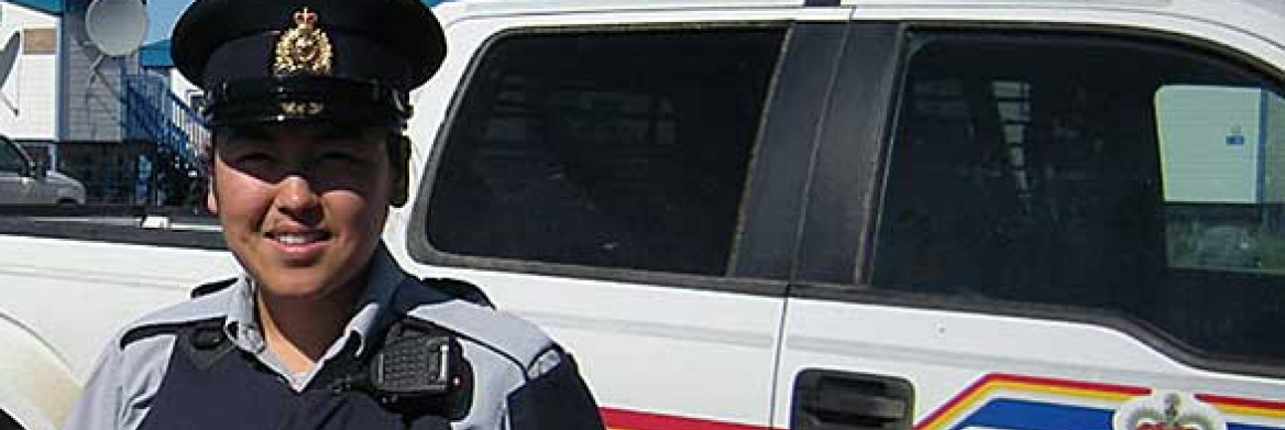 Female aboriginal RCMP officer stands in front of an RCMP cruiser. 