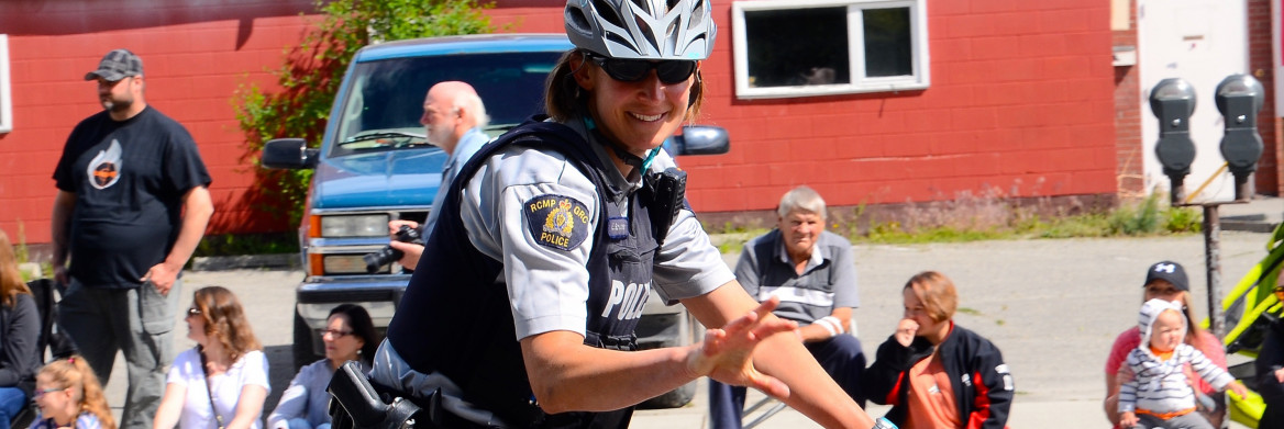 Sous un soleil radieux, montée sur son vélo de travail, une policière de la GRC portant des verres fumés et un casque gris salue les passants qu'elle croise.