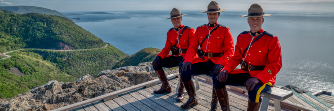 Trois policiers de la GRC en tunique rouge sont assis sur un banc à un point d'observation avec vue sur des collines et l'océan.