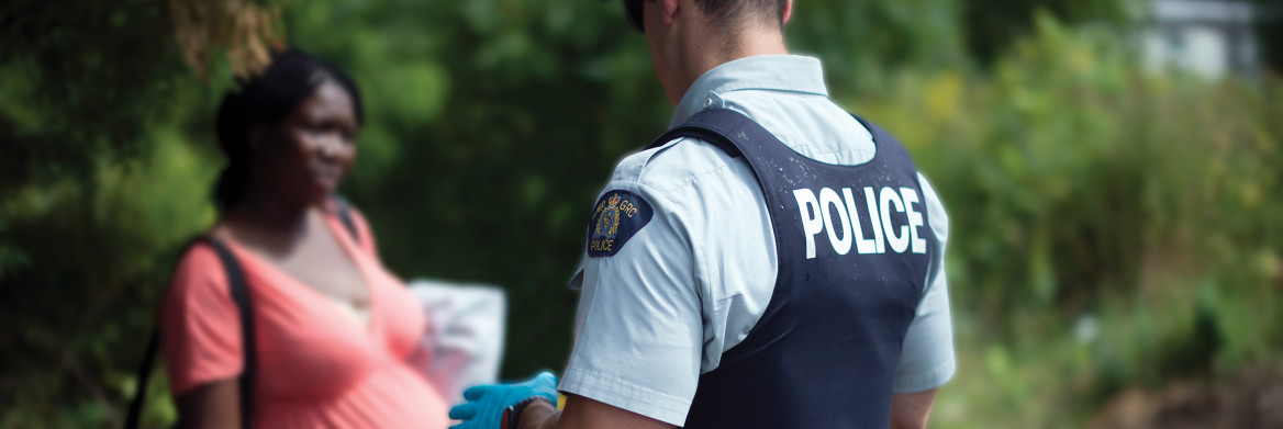 Male police officer speaks to pregnant woman carrying bags in forested area.