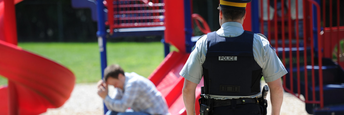 A male police officer approaches a man who is sitting at the bottom of a slide in a playground with his hands against his forehead.