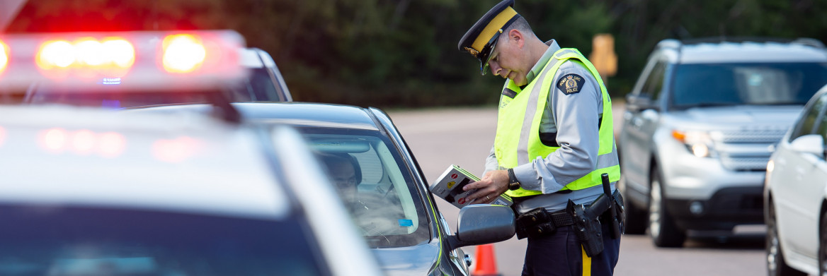 Male police officer wearing a high-visibility vest stands next to a car stopped on a busy highway. He speaks to the male driver while holding a notebook. A police car with lights flashing is parked in the foreground.