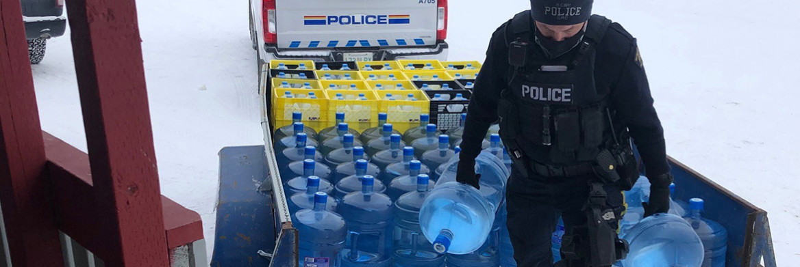 Un policier de la GRC transportant deux grands contenants d'eau. Un camion et une remorque de la GRC en arrière-plan. 