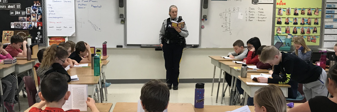 Female police officer holding book stands in front of students in classroom.
