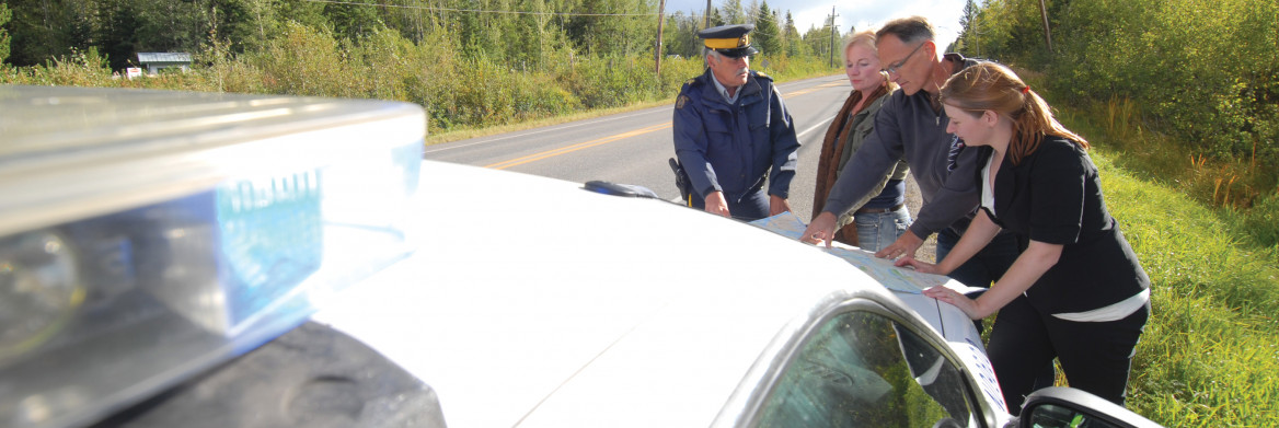 Un policier et trois chercheurs regardent des papiers devant un véhicule de police le long d'une route isolée. 
