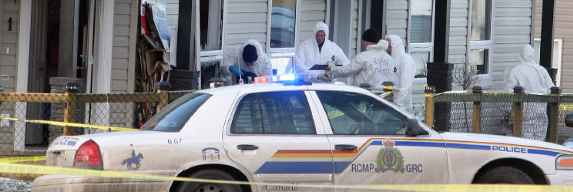 RCMP specialists wearing white protective suits work outside a damaged home. An RCMP cruiser is parked nearby.