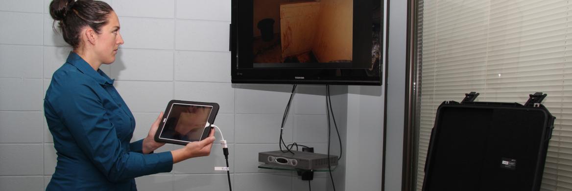 Une femme debout tient une tablette à côté d'un écran de télé.