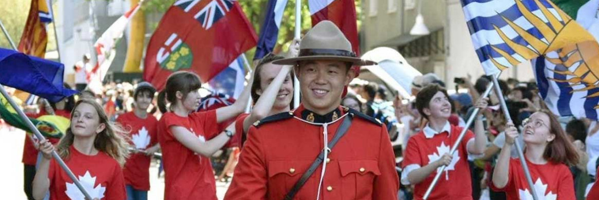 An RCMP officer in red serge walks in a Canada day parade. In the crowd behind him, people wearing red shirts are waving provincial flags.