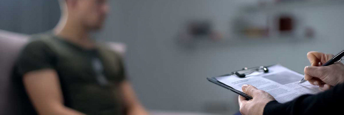 A blurred image of a man sitting facing an unidentified person holding a clipboard and pen in their hand.