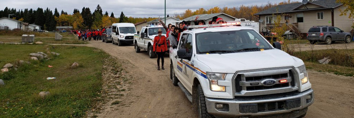A female RCMP officer dressed in red serge stands next to an RCMP truck filled with a group of people who are sitting and waving in the back. On the road behind them are other vehicles and a large crowd of people wearing red.