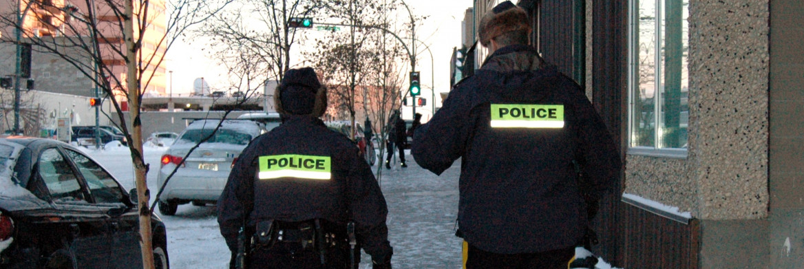 Two police officers walk on snowy sidewalk in city.