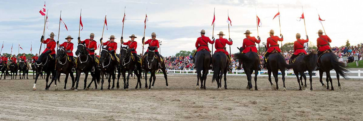 A large group of RCMP members in red serge ride horses while holding the RCMP lance pennon as part of the musical ride with spectators watching on.