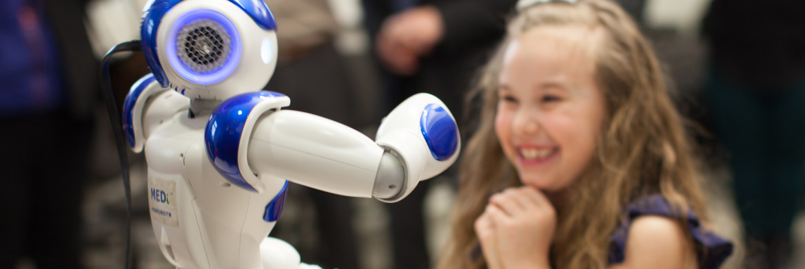 Young girl stands gleefully watching a two-foot-tall white and blue robot standing on a table.