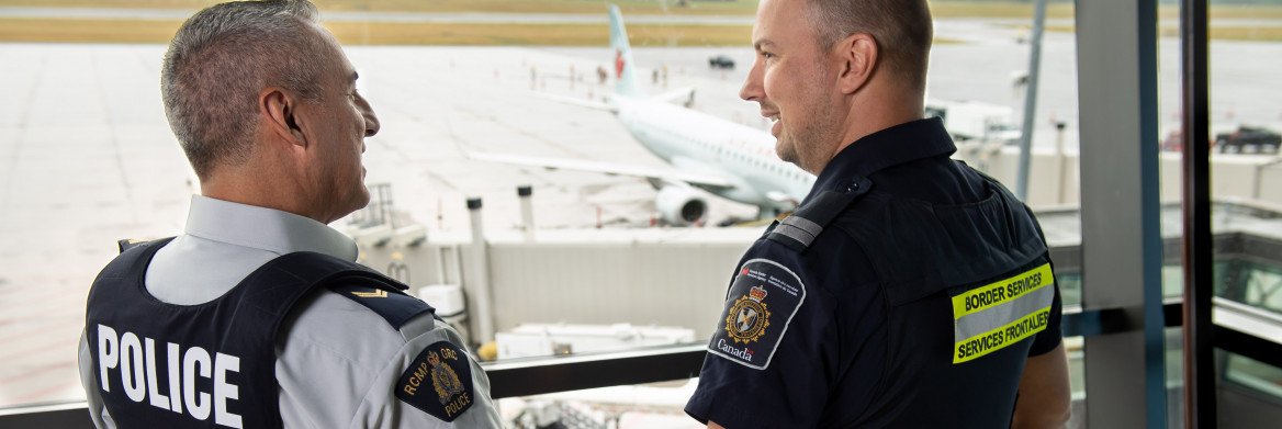 A male RCMP officer and a male CBSA agent talk to each other inside an airport. An airplane can be seen on the tarmac behind them. 