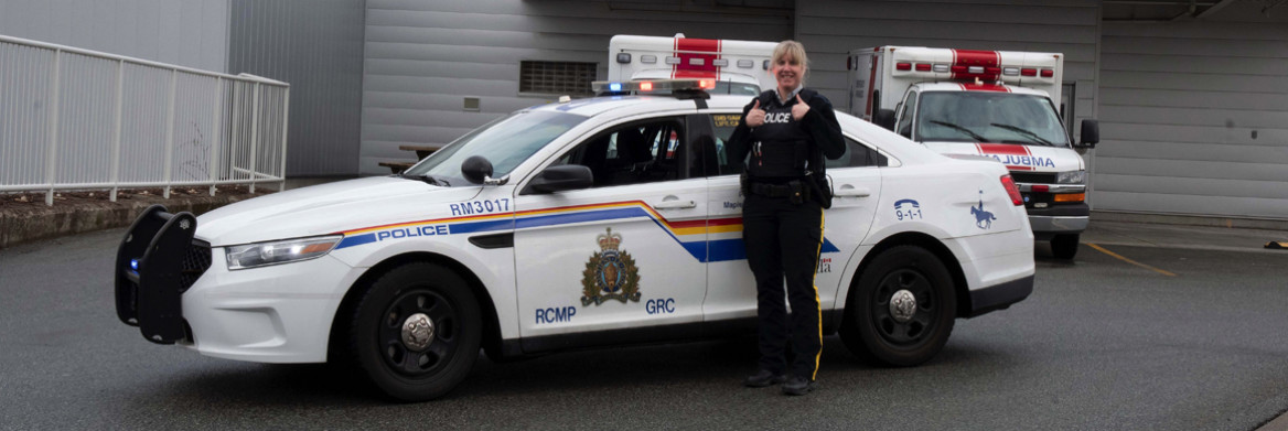 Female RCMP officer giving two thumb's up while standing in front of a police cruiser with a building and ambulances in the background.