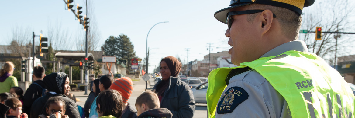 A male police officer stands on a busy intersection near a group of young pedestrians.